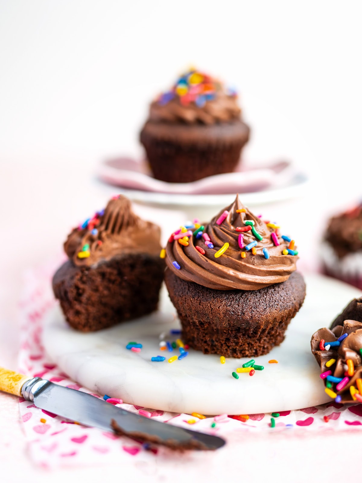 Air fryer chocolate cupcakes with chocolate frosting and rainbow sprinkles sit on a white marble surface; one cupcake is cut in half, showing its moist interior, with a yellow-handled knife nearby.
