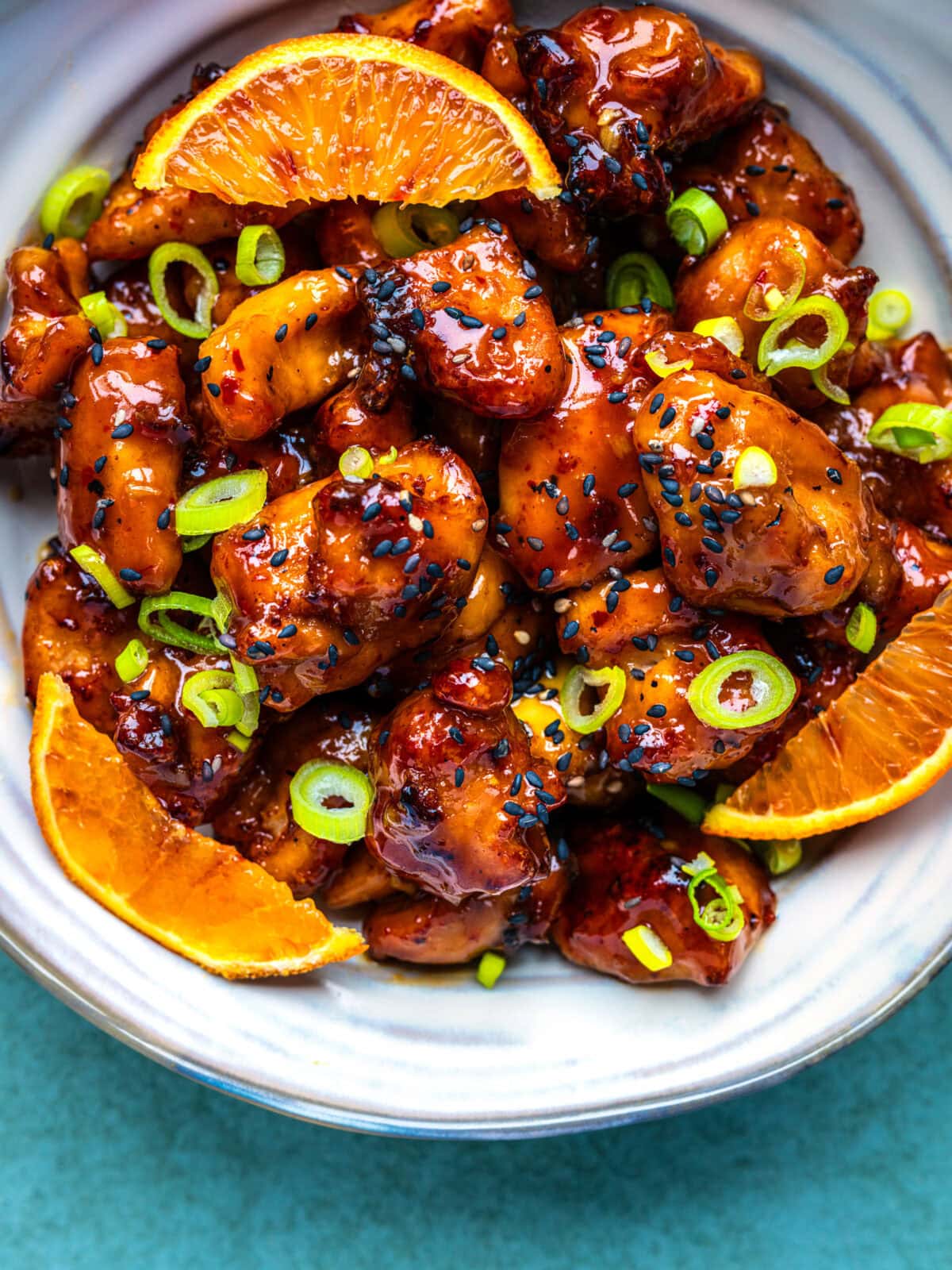 A plate of glazed air fryer orange chicken garnished with sliced green onions, black sesame seeds, and orange wedges, served on a white dish against a blue background.