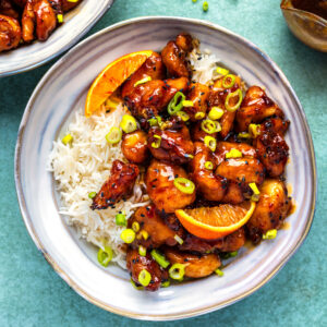 A bowl of white rice topped with glazed air fryer orange chicken pieces, orange slices, and chopped green onions, with extra chicken and sauce in the background on a light green surface.