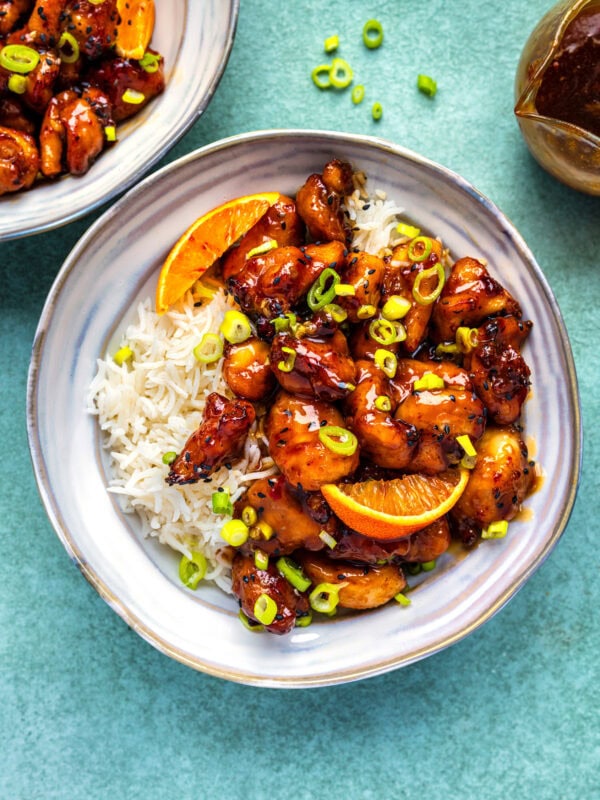 A bowl of white rice topped with glazed air fryer orange chicken pieces, orange slices, and chopped green onions, with extra chicken and sauce in the background on a light green surface.