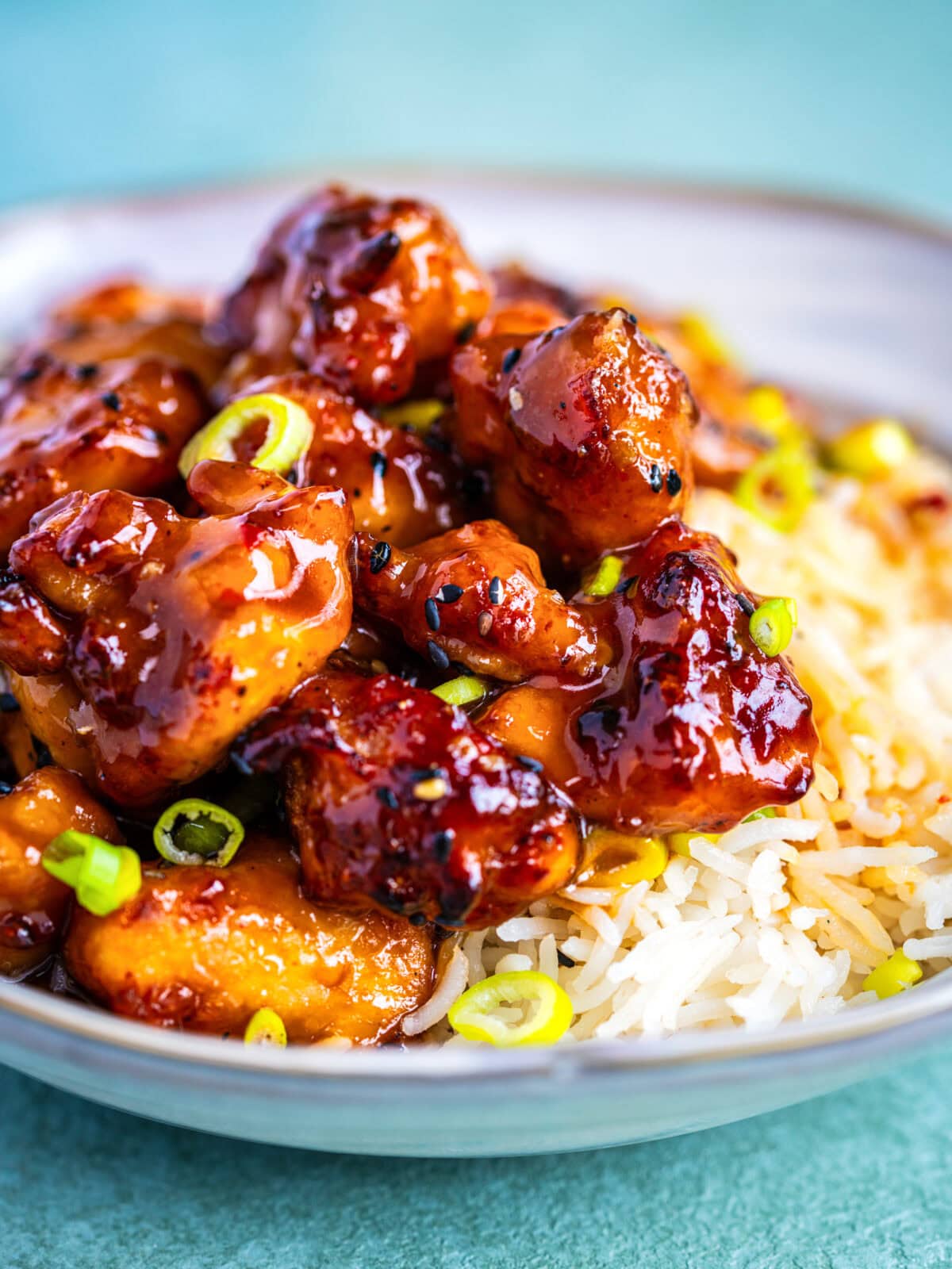 A bowl of white rice topped with glazed, saucy pieces of air fried orange chicken, garnished with chopped green onions, on a light blue surface.