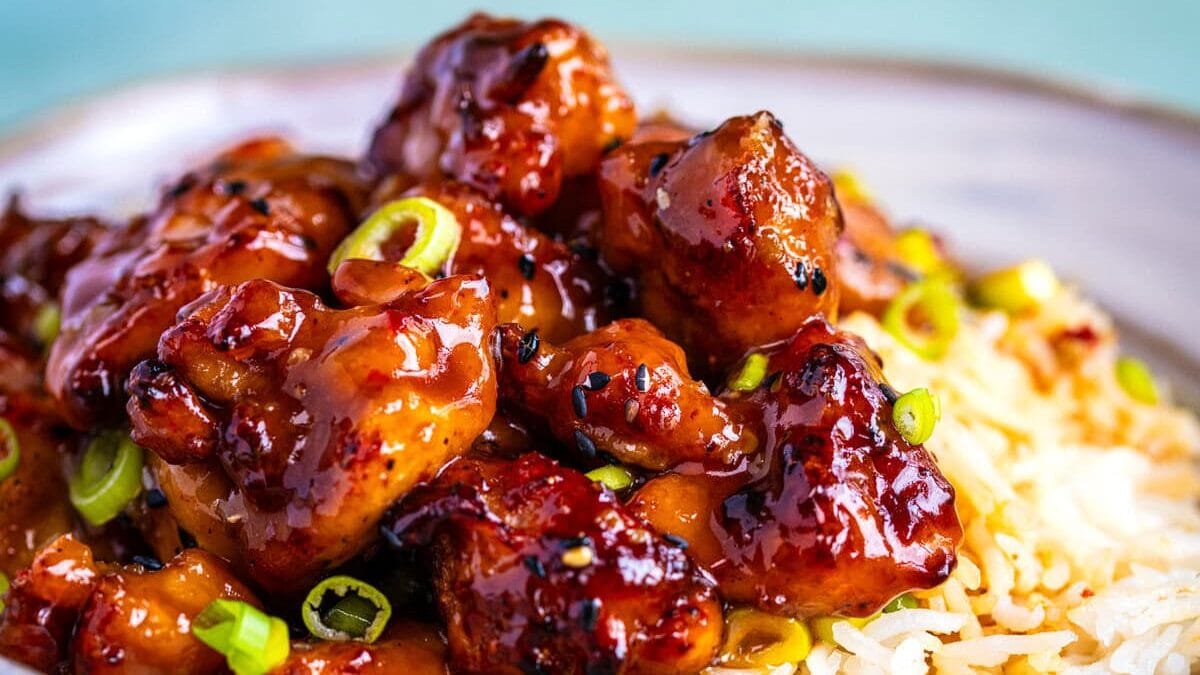 A bowl of white rice topped with glazed, saucy chicken pieces, garnished with sliced green onions, served on a light blue surface.