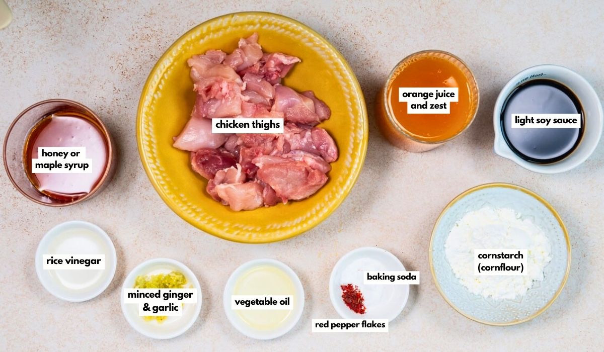 Top-down view of bowls containing ingredients for Air Fryer Orange Chicken: chicken thighs, orange juice and zest, light soy sauce, cornstarch, baking soda, red pepper flakes, vegetable oil, minced ginger and garlic, rice vinegar, and honey or maple syrup.