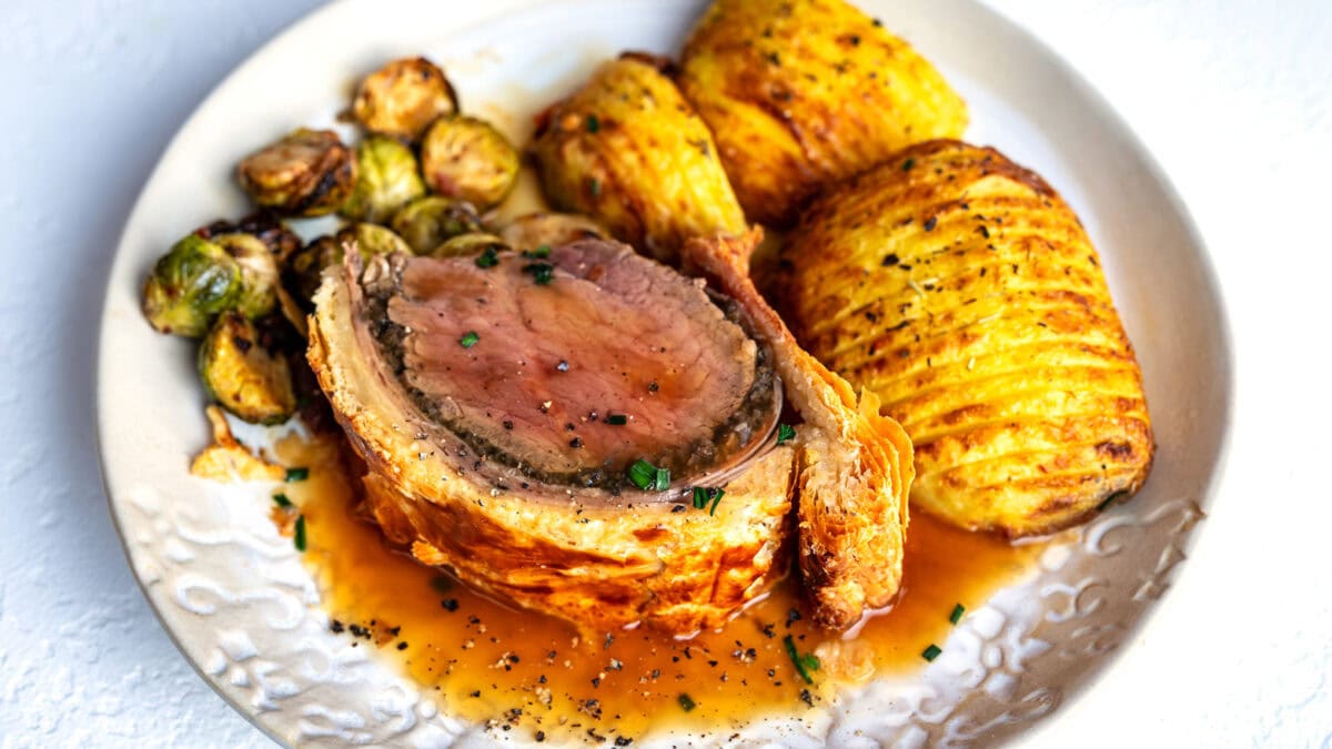 A plate with sliced beef Wellington in pastry, roasted Brussels sprouts, and two golden Hasselback potatoes, topped with sauce and garnished with chopped herbs.