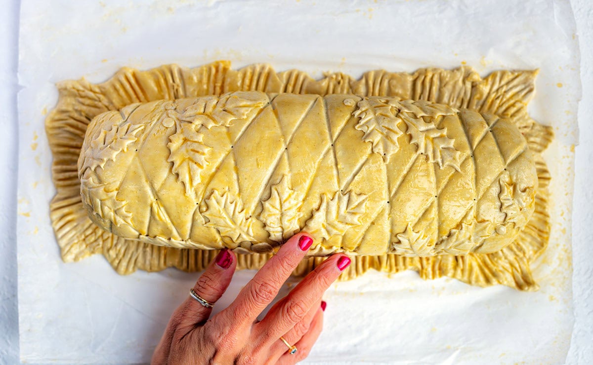 A hand with red-painted nails touches an unbaked Beef Wellington decorated with leaf shapes and a crisscross pattern, resting on parchment paper.