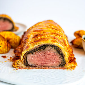 A sliced beef Wellington shows layers of flaky golden pastry, mushroom duxelles, and medium-rare cooked beef tenderloin on a white plate, with more slices and side dishes in the background.