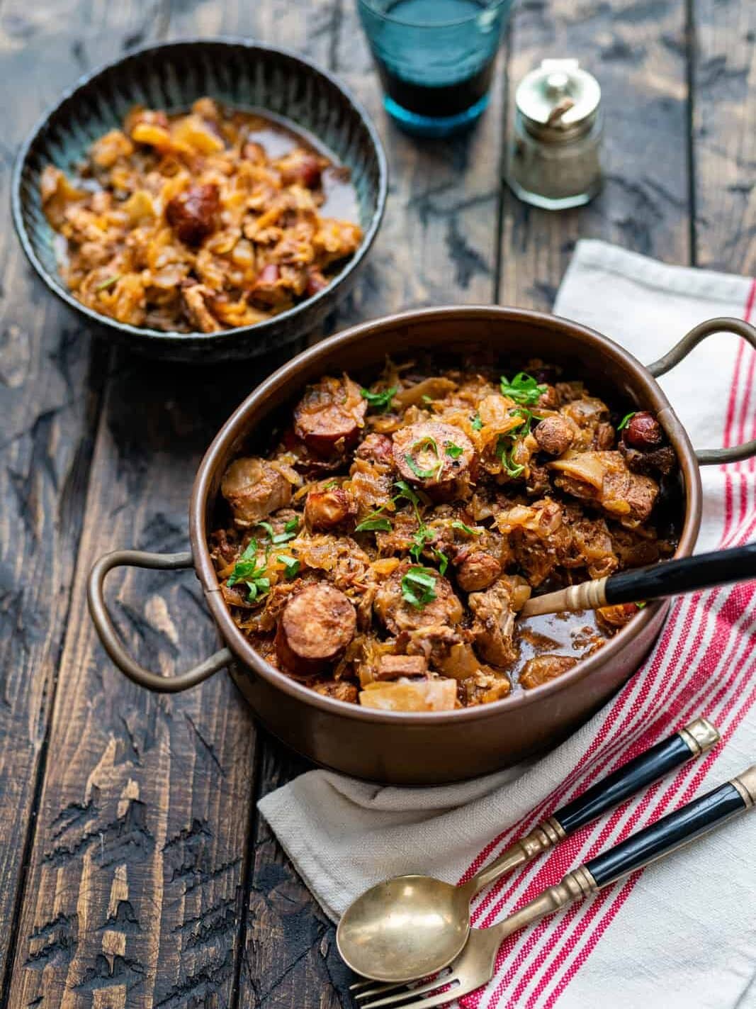 A rustic pot of stew with sausage, meat, and vegetables garnished with herbs sits on a wooden table beside a bowl of the same dish, cutlery, a glass of water, salt and pepper shakers, and a striped napkin.