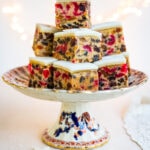 A decorative cake stand holds a stacked arrangement of Christmas Cake Bites topped with white icing, displaying colorful pieces of candied fruit and nuts. Soft lights twinkle in the blurred background.