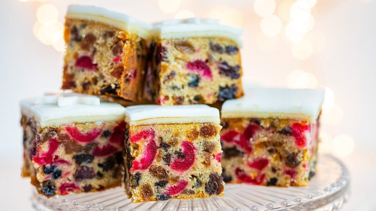 Seven square slices of fruitcake with white icing are displayed on a glass cake stand, with two more slices on plates nearby. The cake is filled with colorful dried fruit. Soft, warm lights blur in the background.