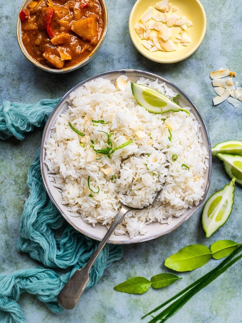 A bowl of coconut rice garnished with lime wedges and sliced almonds, with a spoon resting inside. Nearby are a small bowl of curry, a dish of almonds, lime slices, and herbs on a textured surface.