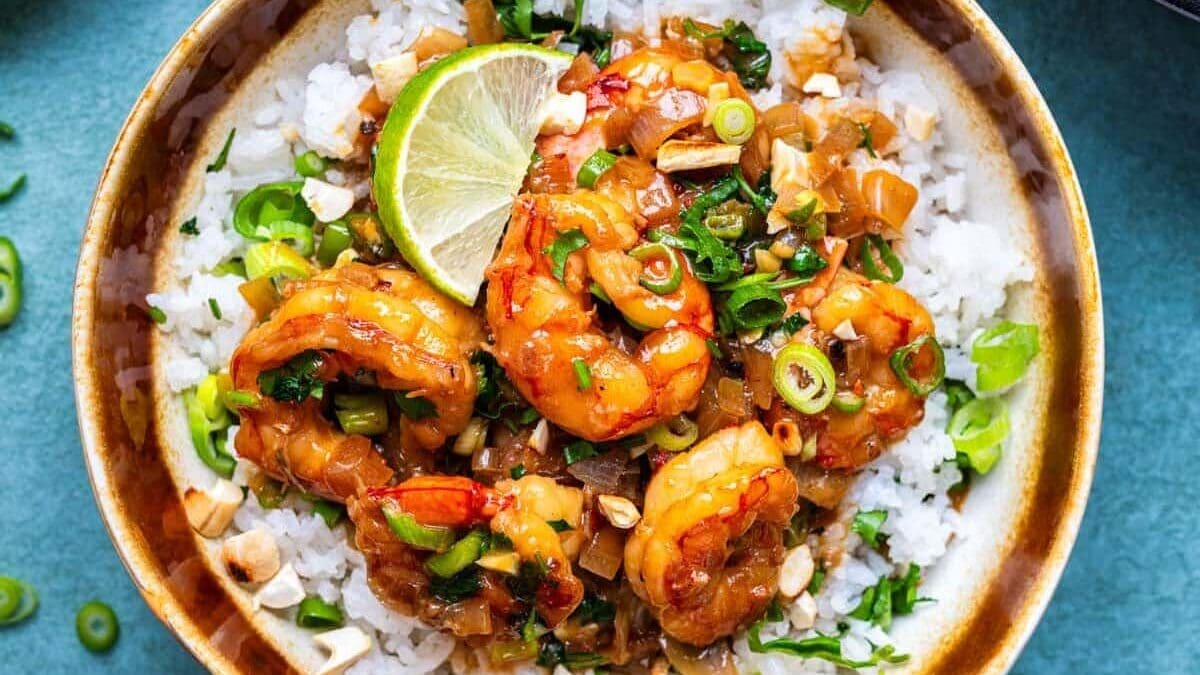 A bowl of coconut jasmine rice topped with saucy coconut shrimp, chopped green onions, cilantro, and a lime wedge. Nearby are bowls of chopped nuts, sliced green onions, and more shrimp in sauce.