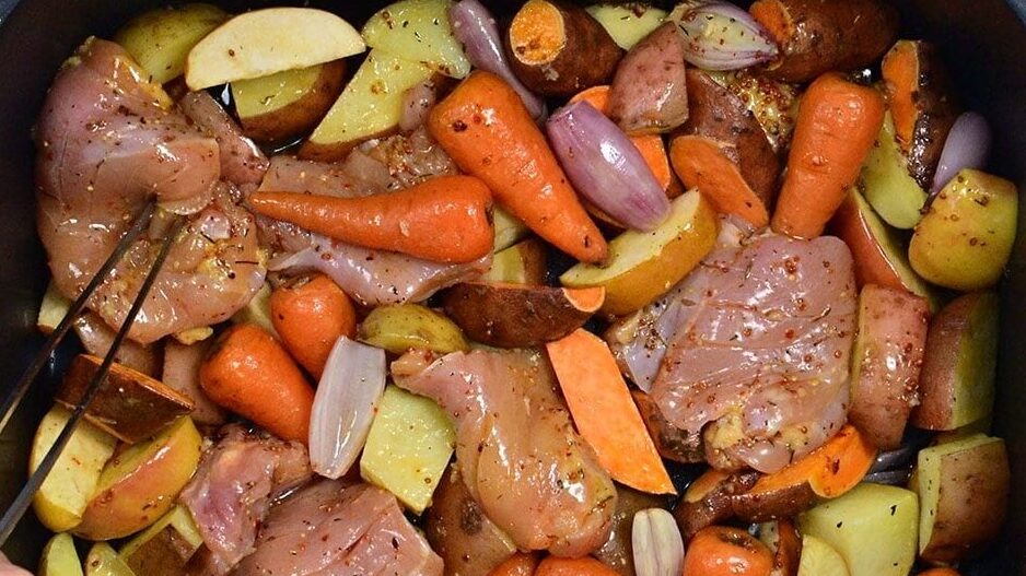 A roasting pan filled with raw chicken thighs, carrots, potatoes, and shallots, seasoned and ready to cook. A hand uses tongs to arrange the ingredients.