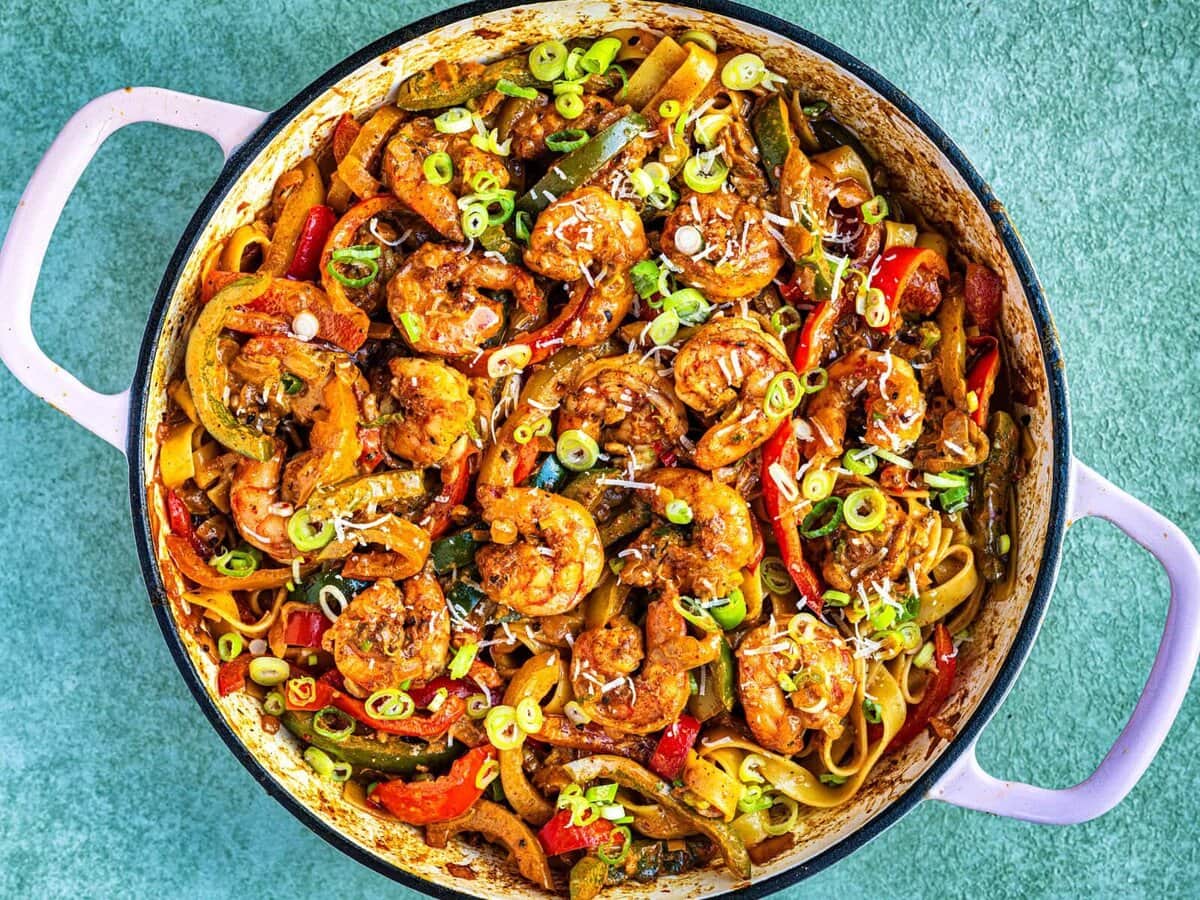 A large pot filled with spicy Rasta pasta, shrimp, bell peppers, and sliced green onions, all coated in a savory sauce. The dish is colorful with red, yellow, and green vegetables, and the pot sits on a light green textured surface.