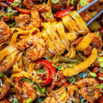 A close-up of rasta pasta with jerk shrimp with wide noodles, shrimp, sliced red peppers, green onions, and a sprinkle of red pepper flakes, garnished with grated cheese and herbs.