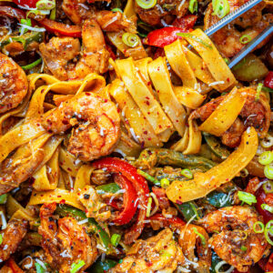 A close-up of rasta pasta with jerk shrimp with wide noodles, shrimp, sliced red peppers, green onions, and a sprinkle of red pepper flakes, garnished with grated cheese and herbs.