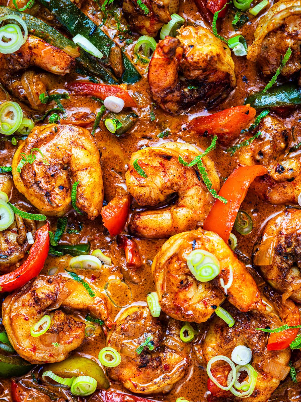Close-up of spicy shrimp cooked in a rich, reddish-brown sauce with sliced green onions and red and green bell peppers, garnished with herbs. The dish looks flavorful and vibrant.
