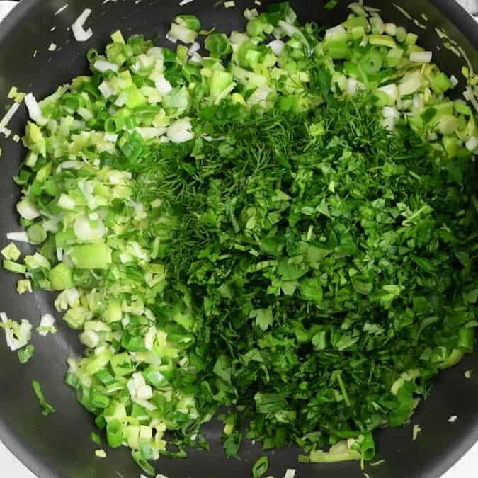 Chopped green onions, parsley, and dill cooking in a black skillet, with the herbs and onions spread evenly across the surface.