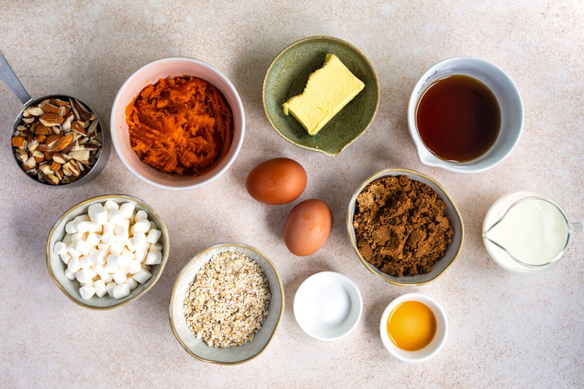 A top view of various baking ingredients in bowls, including chopped nuts, pumpkin puree, butter, maple syrup, eggs, brown sugar, cream, marshmallows, oats, salt, and vanilla extract, arranged on a light surface.