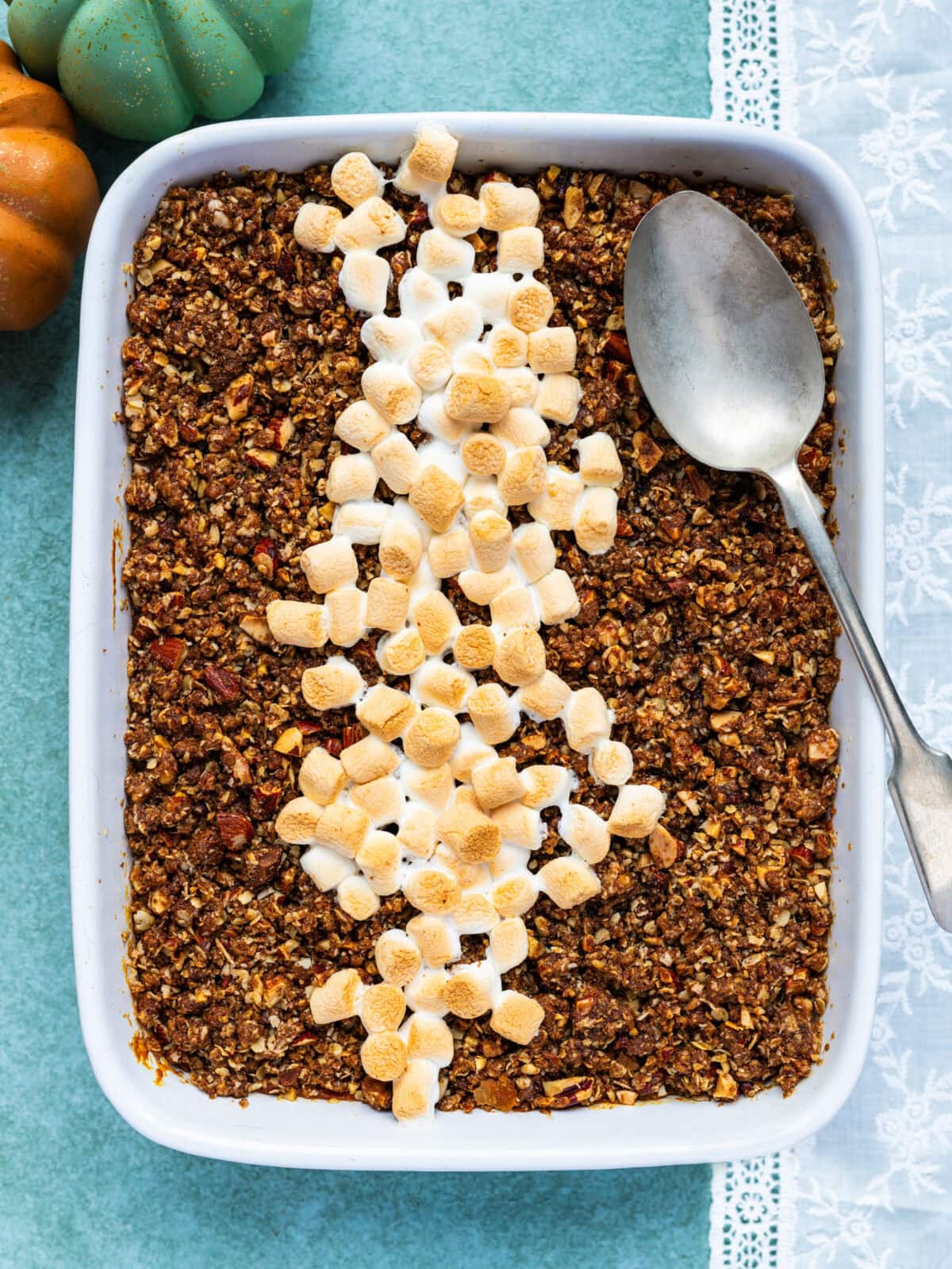 A baked sweet potato casserole topped with a brown crumbly layer and a line of toasted mini marshmallows in the center, with a large serving spoon resting on one side. Two small decorative pumpkins are nearby.