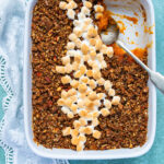 A rectangular baking dish filled with sweet potato souffle casserole, topped with pecan crumble and a diagonal line of toasted mini marshmallows. A serving has been scooped out, with a spoon resting in the dish.