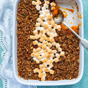 A rectangular baking dish filled with sweet potato souffle casserole, topped with pecan crumble and a diagonal line of toasted mini marshmallows. A serving has been scooped out, with a spoon resting in the dish.