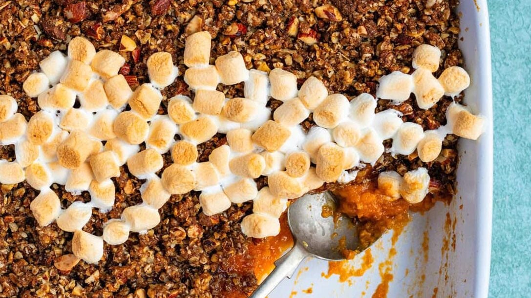 A rectangular baking dish filled with sweet potato souffle casserole, topped with pecan crumble and a diagonal line of toasted mini marshmallows. A serving has been scooped out, with a spoon resting in the dish.