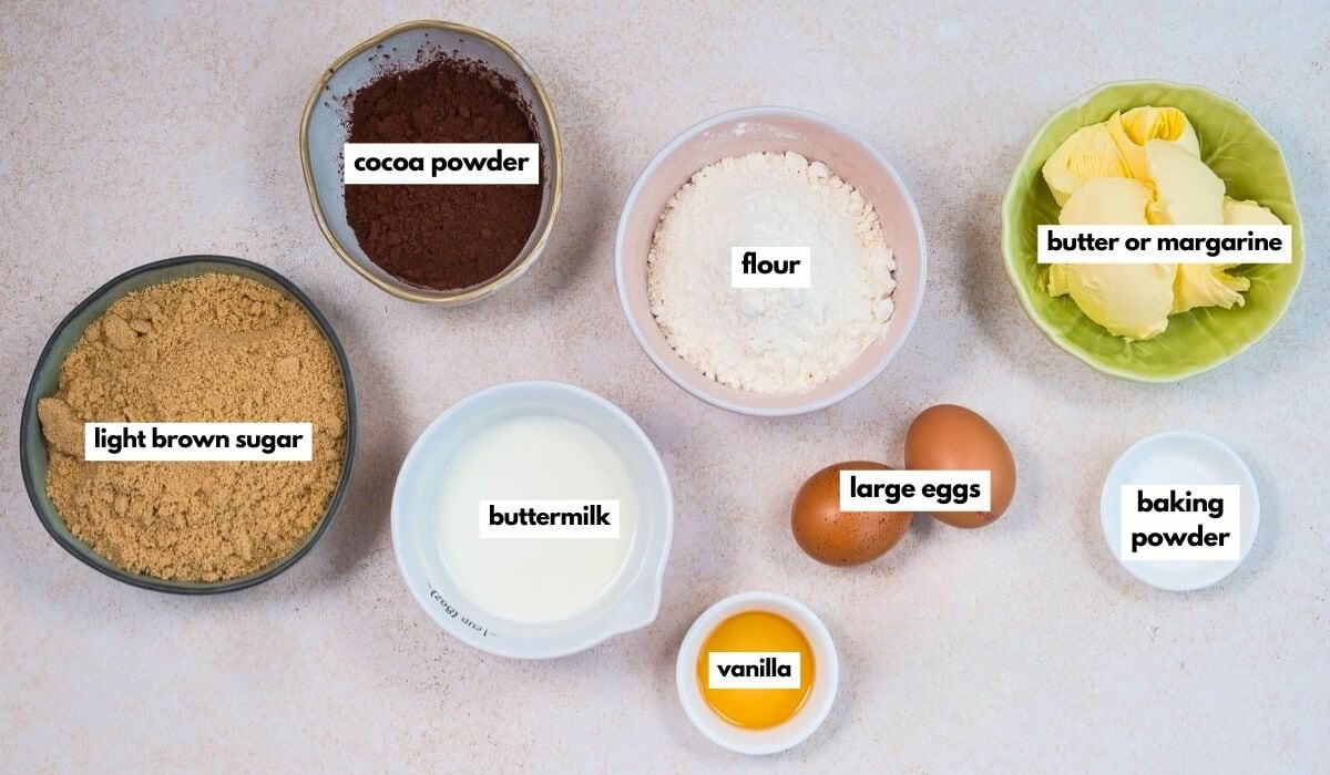 Top-down view of baking ingredients in bowls: cocoa powder, flour, butter or margarine, light brown sugar, buttermilk, vanilla, large eggs, and baking powder, each labeled with white text on the image.