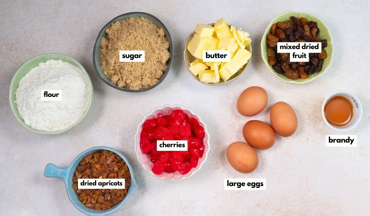Ingredients for baking Mary Berry Christmas cake bites are displayed in bowls on a white surface: flour, brown sugar, butter, mixed dried fruit, brandy, large eggs, dried apricots, and glacé cherries—each bowl neatly labeled.
