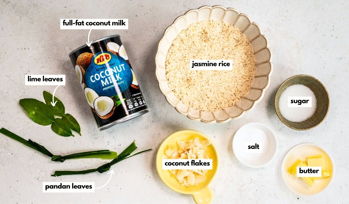 A can of coconut milk, a bowl of jasmine rice, small bowls of sugar, salt, and butter, a dish of coconut flakes, two pandan leaves, and two lime leaves are arranged on a light surface. Each item is labeled.