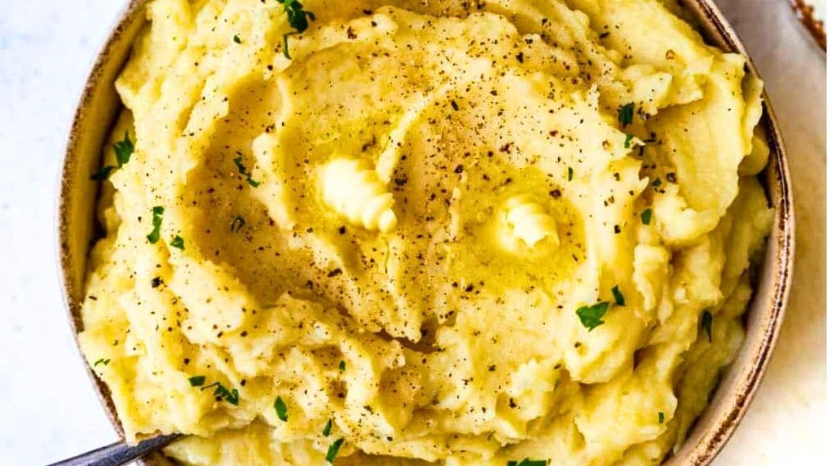 A bowl of creamy Slow Cooker Mashed Potatoes topped with two small pats of butter, freshly ground black pepper, and garnished with chopped parsley. A spoon rests on the side of the bowl.