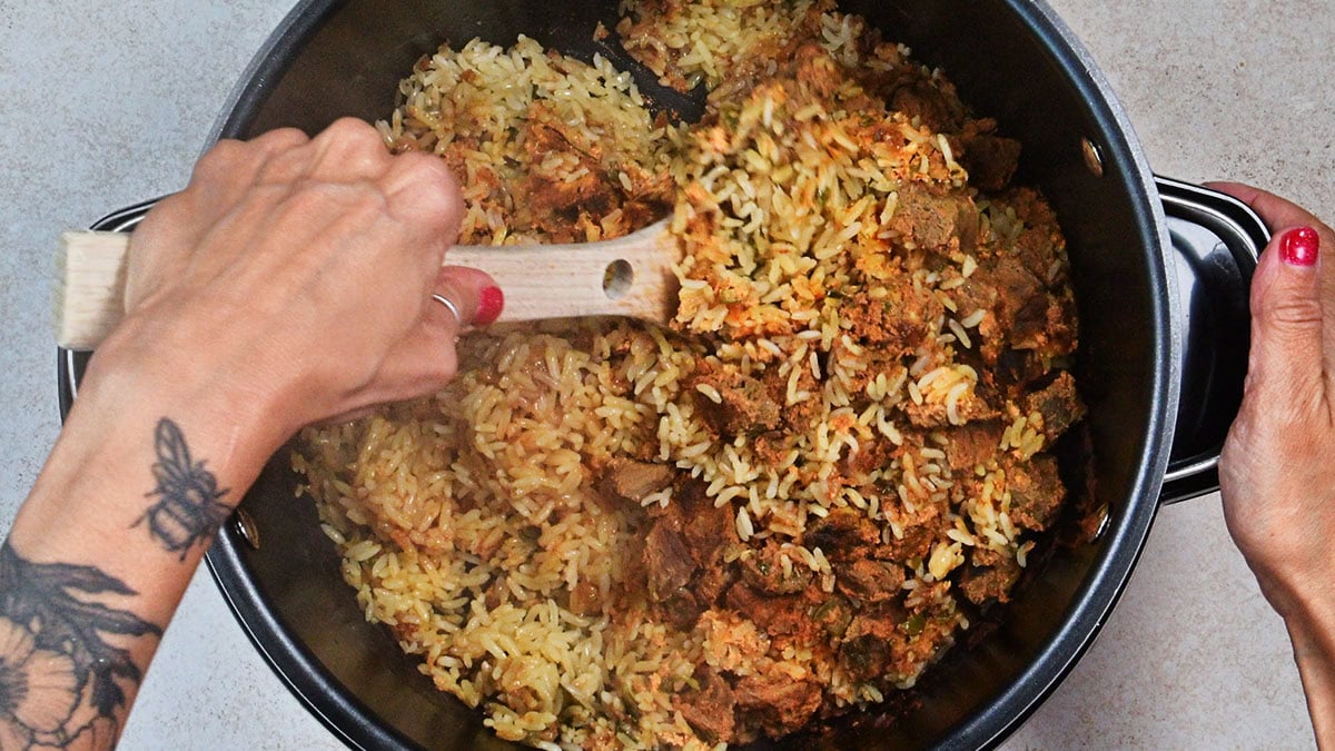 A person with a floral tattoo on their forearm stirs a pot of beef biryani cooked in crockpot with a wooden spoon. The pot sits on a light-colored countertop, and their fingernails are painted red.
