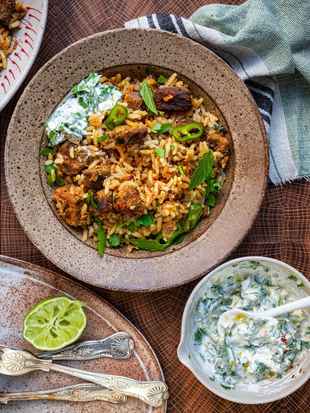 A bowl of beef biryani with spiced rice, grilled meat, sliced green chili, and mint leaves, served alongside creamy herb yogurt sauce and a halved lime on a wooden surface. A striped cloth napkin and cutlery are nearby.