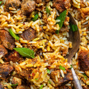 Close-up of a flavorful crockpot beef biryani with chunks of spiced meat, garnished with fresh green herbs. A spoon is partially visible, scooping up the rice and meat mixture.