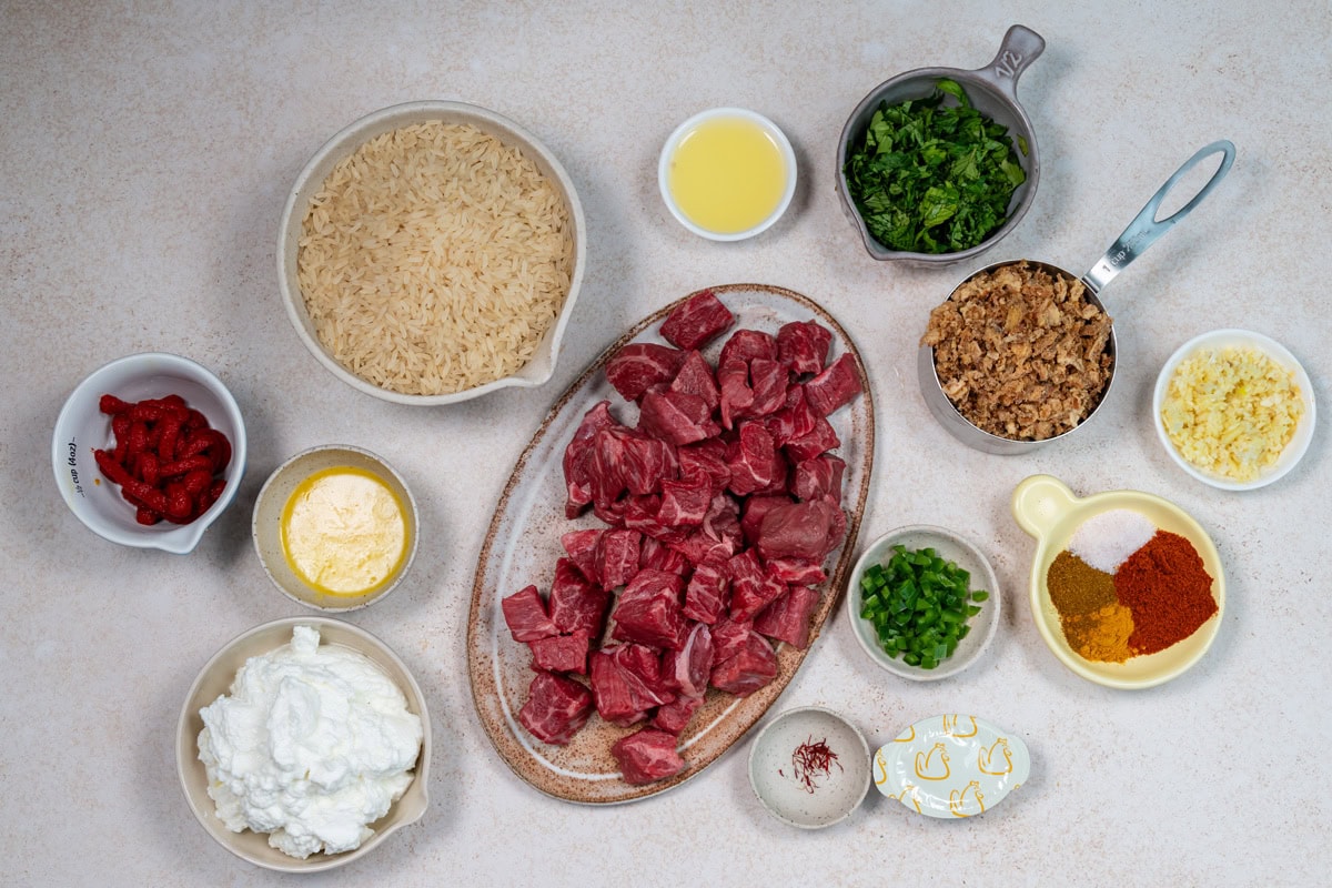 An overhead view of raw ingredients for a dish, including cubed beef, rice, chopped herbs, dried onions, minced garlic, yogurt, butter, peppers, spices, tomato paste, and lemon juice arranged on a light countertop.