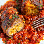 Two slices of beef braciole stuffed with breadcrumbs and herbs, topped with chopped parsley, served on a plate with chunky tomato sauce and a fork beside the food.