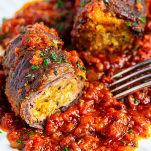 Two slices of beef braciole stuffed with breadcrumbs and herbs, topped with chopped parsley, served on a plate with chunky tomato sauce and a fork beside the food.