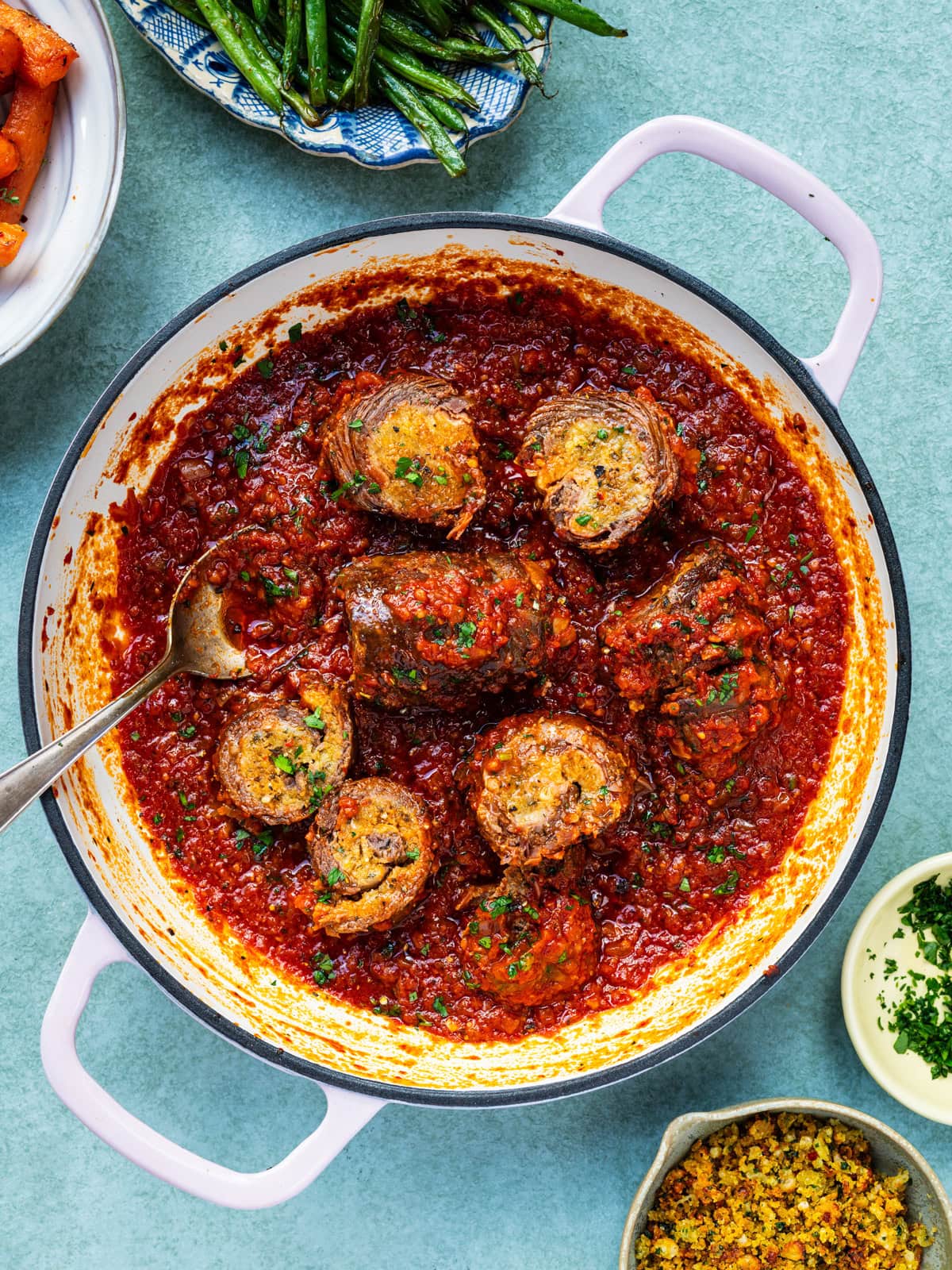 A pan of beef roulades topped with chopped herbs, served in a rich tomato sauce. A spoon rests in the pan. Surrounding the pan are bowls of green beans, carrots, and a small dish of breadcrumbs and herbs.