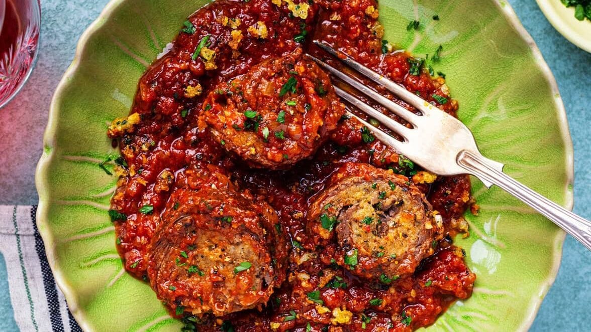 A green plate with three slices of beef braciole covered in chunky tomato sauce, garnished with herbs, with a fork resting on the plate. A striped napkin and sides, including green beans, are partially visible nearby.