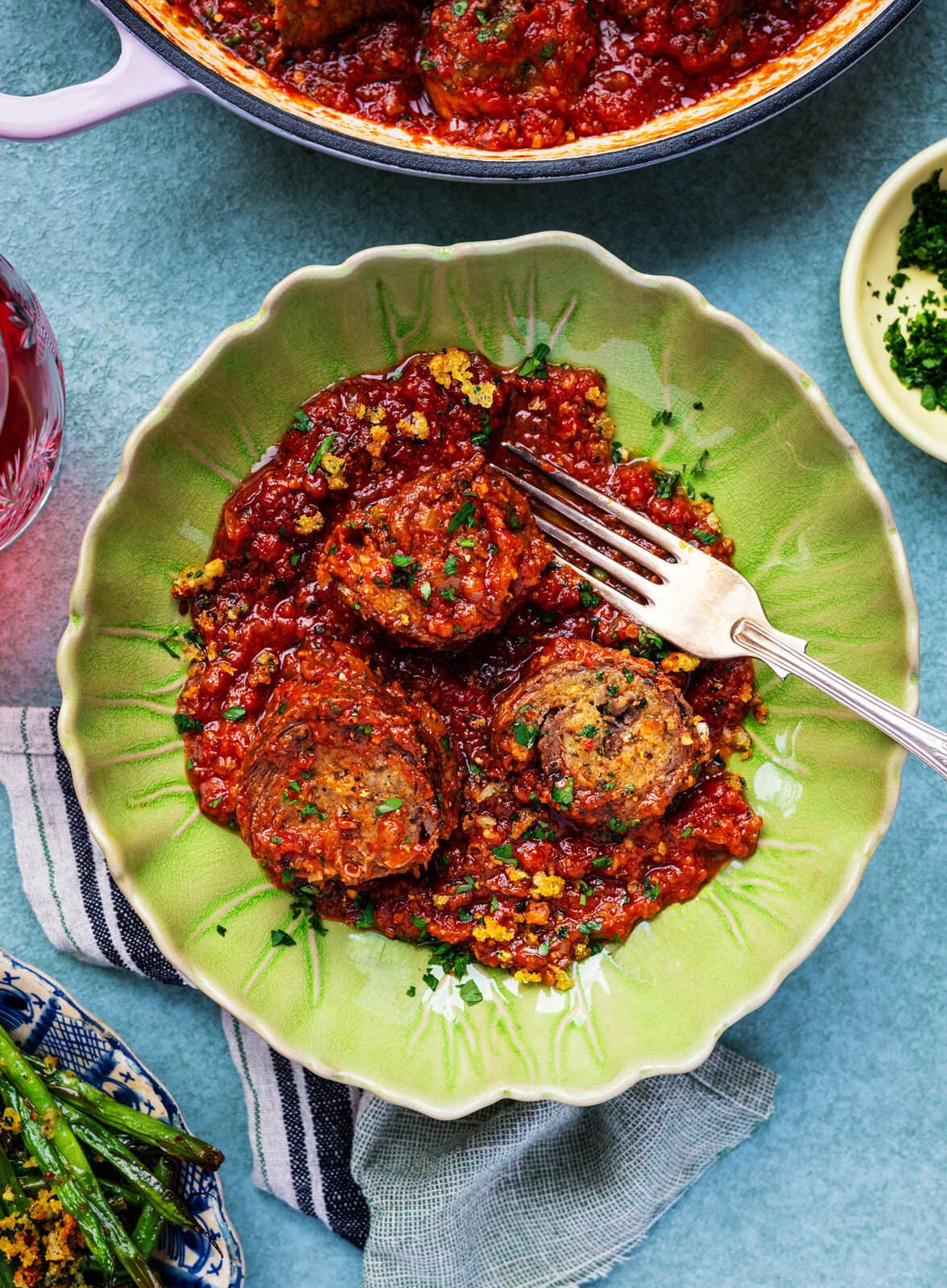 A green plate with three slices of beef braciole covered in chunky tomato sauce, garnished with herbs, with a fork resting on the plate. A striped napkin and sides, including green beans, are partially visible nearby.