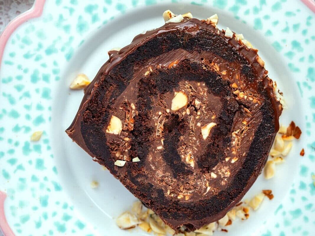 A slice of chocolate Swiss roll with chopped nuts on top sits on a decorative blue and white plate, placed on a lacy white tablecloth. Other colorful plates are partially visible in the background.
