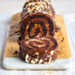 A chocolate Brownie Yule log with a creamy filling, topped with chopped nuts and drizzled chocolate, sits sliced on a wooden board with a marble base.