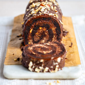 A chocolate Brownie Yule log with a creamy filling, topped with chopped nuts and drizzled chocolate, sits sliced on a wooden board with a marble base.