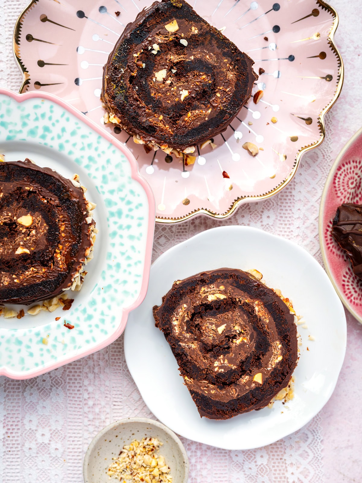 Three slices of chocolate brownie roll topped with chopped nuts are served on decorative plates, set on a lace tablecloth with a small bowl of extra nuts nearby.