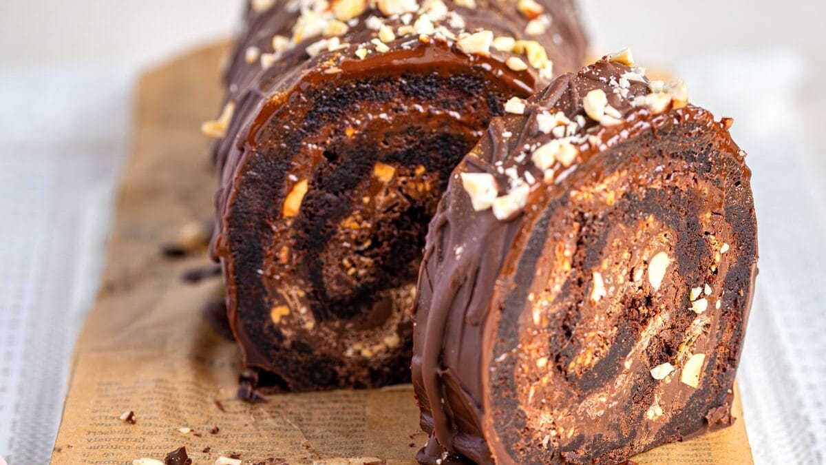 A chocolate brownie roll is topped with chopped nuts and sliced on a parchment-lined marble board. The cake features a rich swirl pattern inside and is elegantly drizzled with chocolate.