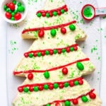 A Christmas tree-shaped cake with white frosting, decorated with red icing, green sugar sprinkles, and red and green candies, sits on a white platter. A small bowl of candies and a teaspoon of sprinkles are nearby.