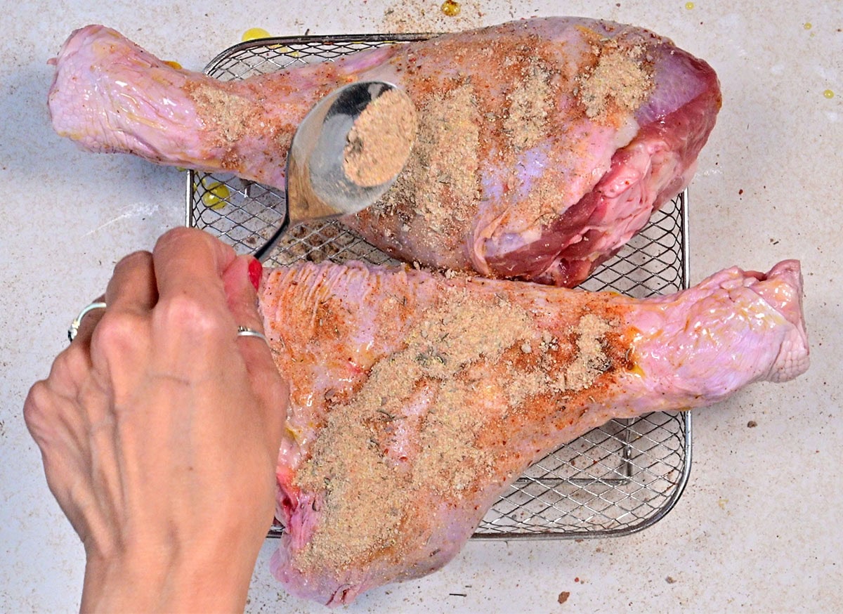 A hand sprinkles seasoning onto two large, raw turkey legs placed on a wire rack, preparing them for cooking.