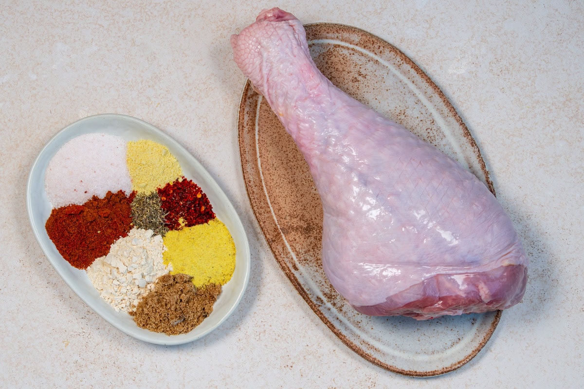 A raw turkey leg on an oval plate sits beside a dish containing piles of various spices and seasonings, including yellow, red, white, and brown powders, on a light-colored surface.