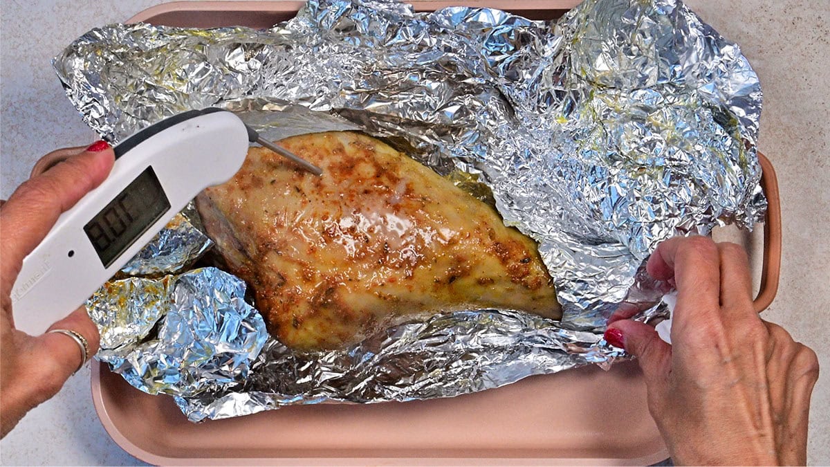 A person checks the temperature of cooked chicken breast wrapped in aluminum foil using a digital meat thermometer. The chicken rests in a pink baking tray on a light countertop.