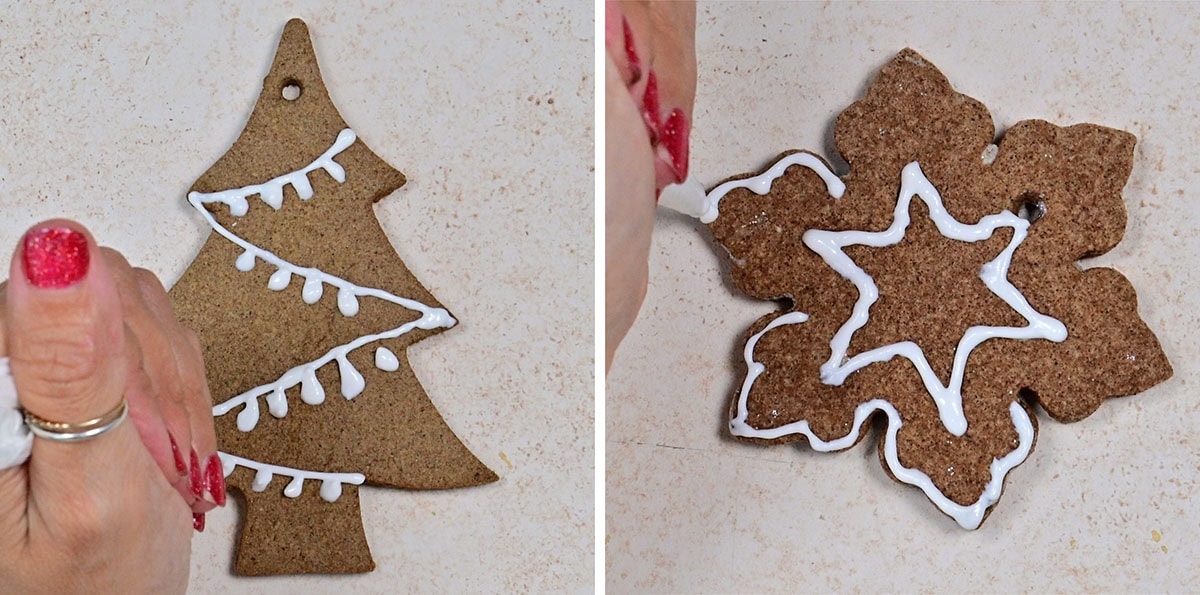 Two hands decorate Christmas gingerbread ornaments with white icing; one is a Christmas tree with garland, the other is a snowflake with star and outline patterns. Both cookies are on a light countertop.