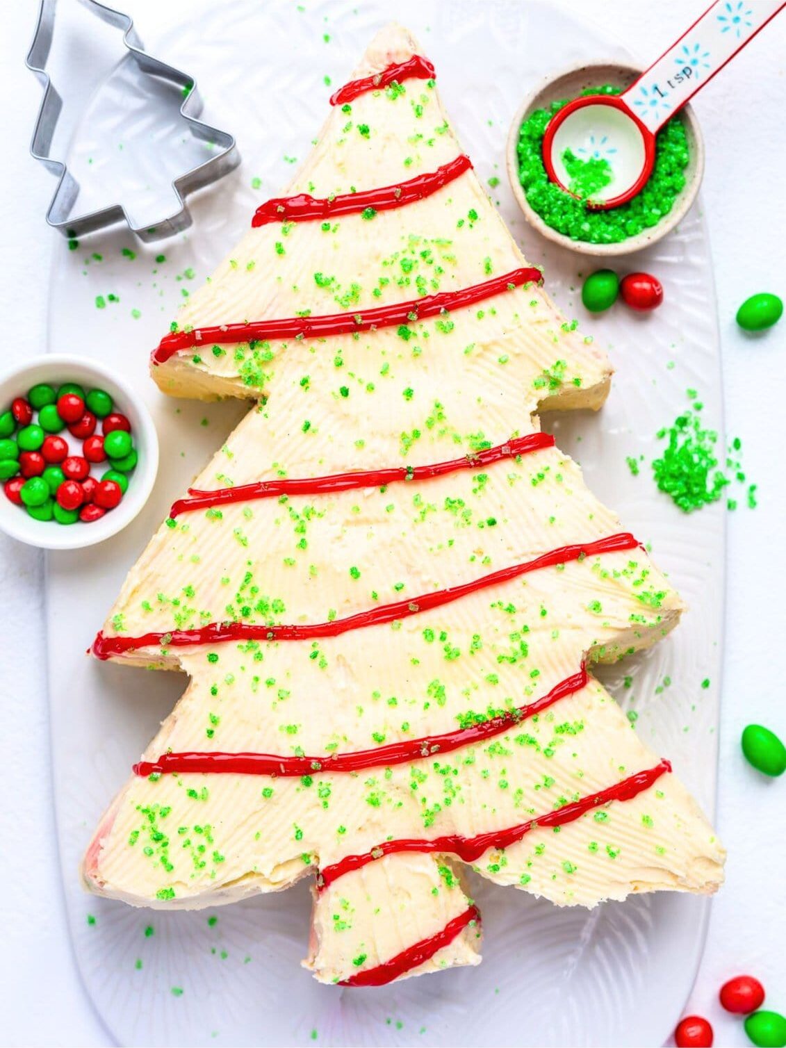 A festive Christmas Tree Cake with white frosting, red icing garlands, and green sprinkles sits on a white platter. Red and green candies, a measuring spoon, and a tree-shaped cookie cutter surround this delightful holiday centerpiece.